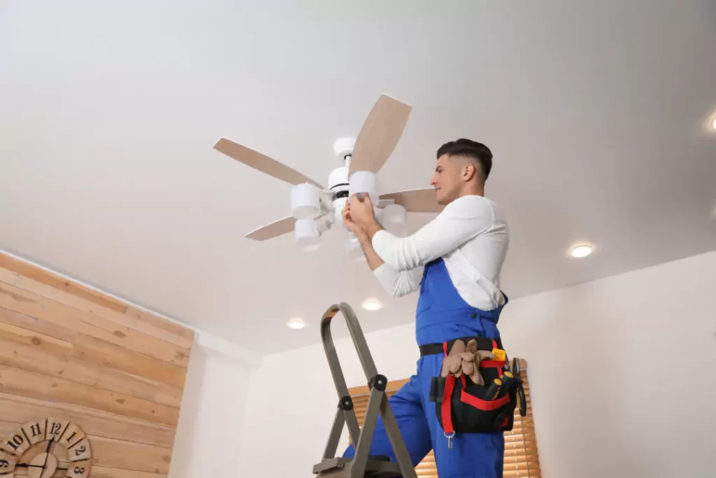 Electrician on ladder installing a white ceiling fan with wooden blades in a modern room.