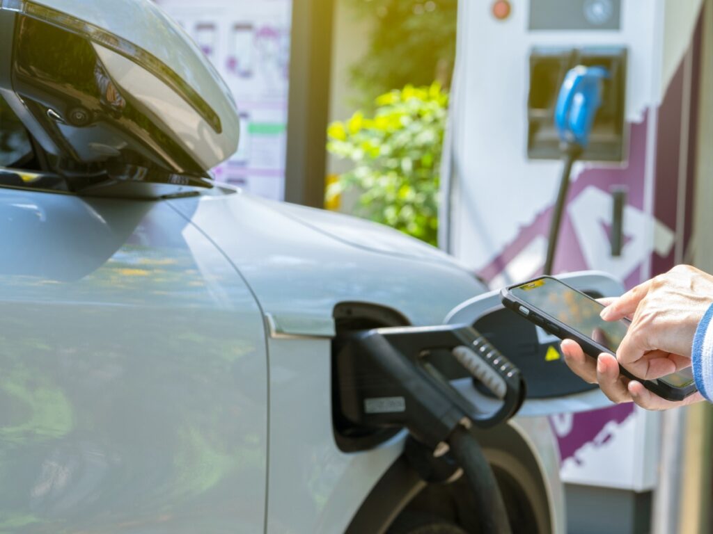 Professional EV charger installation by Sunshine Coast Electrician - person using mobile phone while charging electric vehicle at outdoor charging station, showcasing home electric vehicle charging solutions and EV charging station setup services
