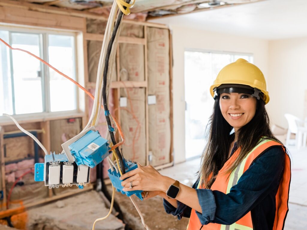 Professional Sunshine Coast Electrician performing house rewiring services - woman in hard hat and safety vest working on exposed electrical wiring during safe electrical upgrades in partially constructed room with insulation and wall framing